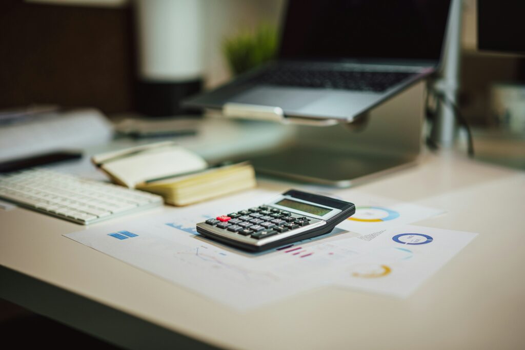 A calculator on printed papers next to an open notebook and a laptop